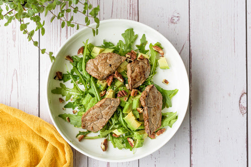Biefstuksalade met rucola, avocado blokjes en pecannoten in een witte kom op een houten tafel — koolhydraatarm keto lunchrecept