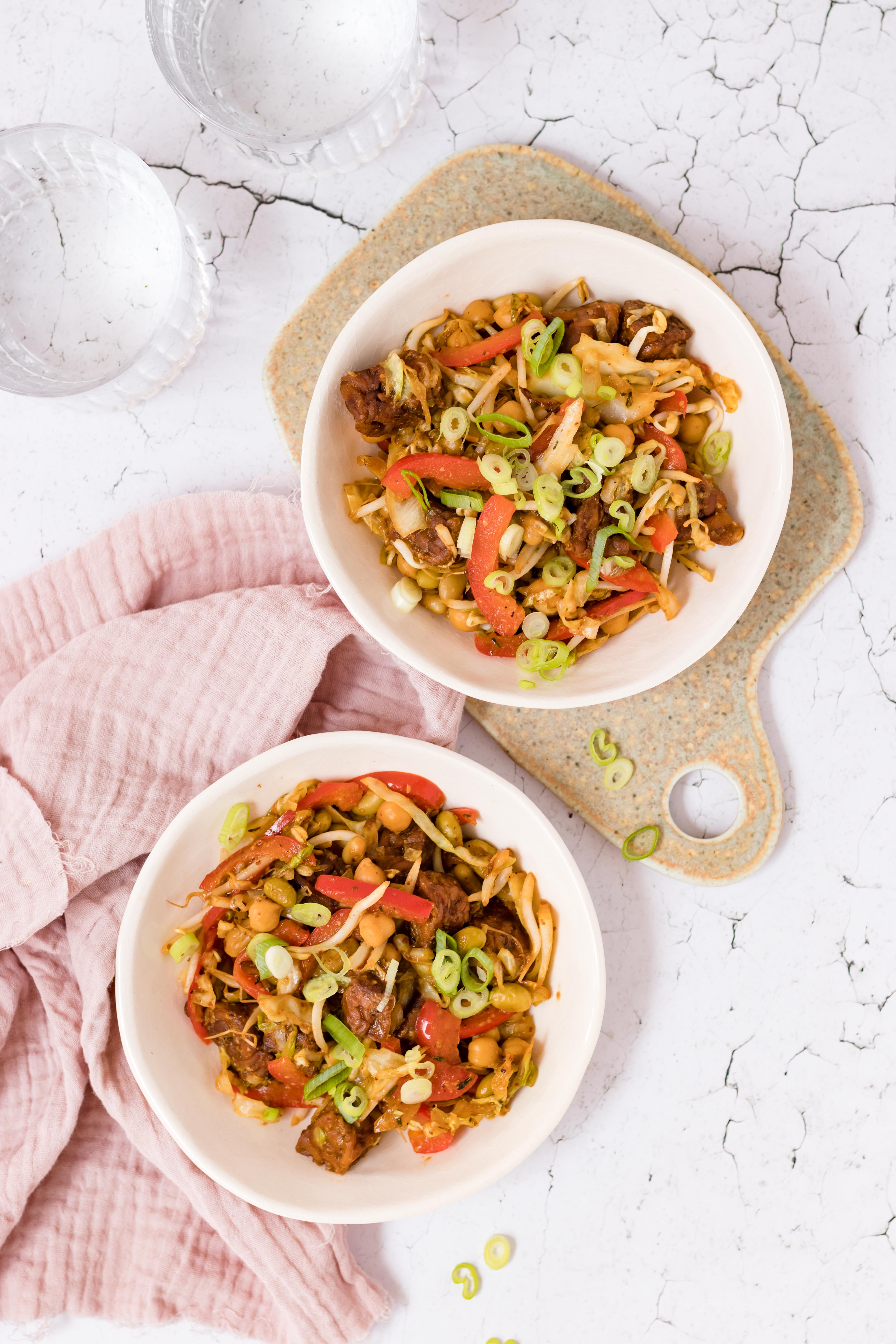 Chinese veggie wok met tempeh, edamame, taugé en rode paprika in twee witte kommen, gegarneerd met bosui en sesamzaad