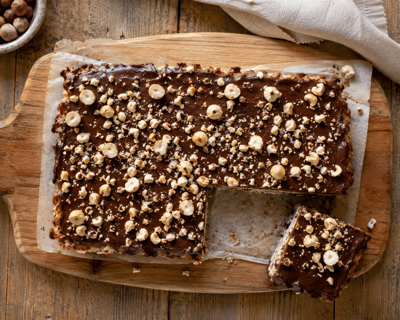 Koolhydraatarm hazelnoot gebak met pure chocolade topping en gehakte hazelnoten, bovenaanzicht op een houten snijplank met één stuk apart