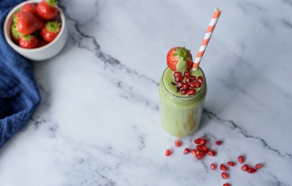 Granaatappelsmoothie in een glazen pot met rietje, gegarneerd met granaatappelpitjes en aardbei, op een marmeren ondergrond